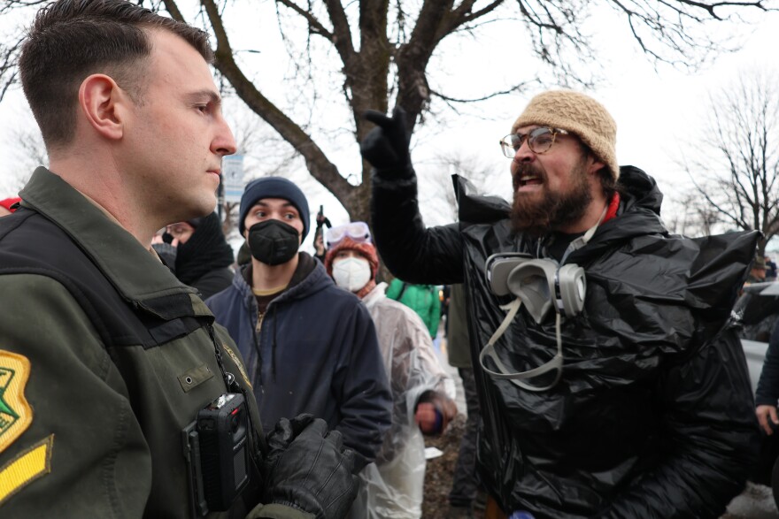 A man in a black garbage bag and wearing a respirator mask around his neck yells angrily at a uniformed officer.