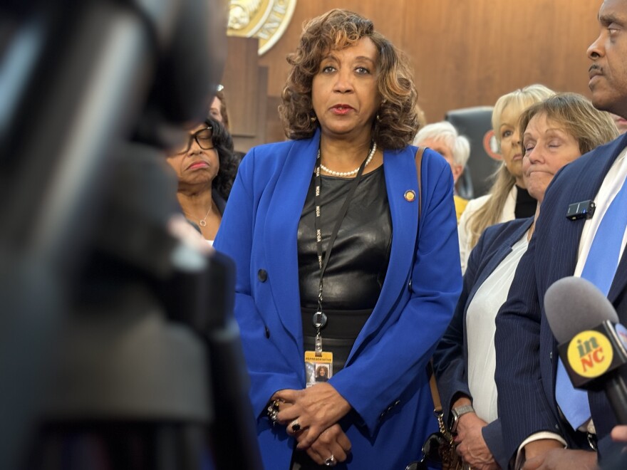 A woman stands with her hands clasped facing cameras and microphones in the foreground. She was shoulder-length hair and is wearing a black shirt and blue blouse.