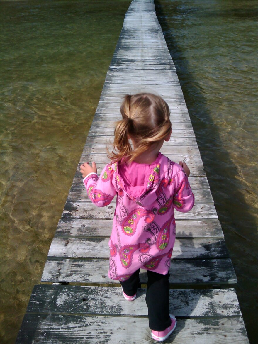 A little girl with blond ponytails and ap ink hooded coat walks on a long pier stretching into a body of water.
