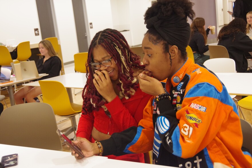 WMU roommates laughing at a TikTok video in the Student Center.  On the left is Blessing Nwamgbe in a red two-piece outfit. She holds her hand to her face as she laughs.  Her hair has bright red and blonde streaks and curls at the ends.  Angelina Opoku-Diawuo is wearing a bright orange, bright blue and black ATT Nascar jacket, covered in patches.  She wears her hair in a topknot, partially wrapped with a dark band.  Her hand is also close to her mouth as she reacts to the video.