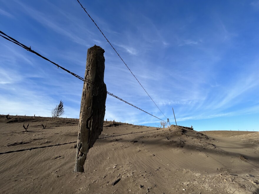 A half-burned wooden fence post hangs on barbed wire near Koester's ranch in Russell County. The wind covered the burned grass in this area with a layer of tan soot and dust.-David Condos/Kansas News Service