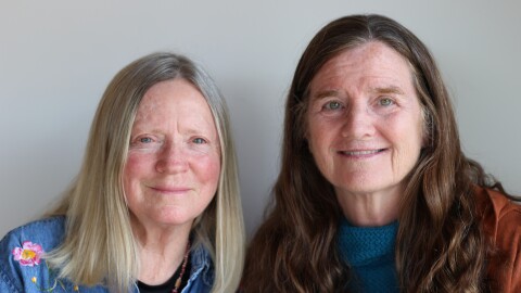 Kathleen Kavarra-Corr and Bernadette Kane-Cole side by side, smile into the camera.  Dr. Kavarra-Corr has straight, shoulder length blonde hair, parted in the middle.  She wears a blue denim  machine embroidered button-up shirt with floral designs, partially unbuttoned over a black tee shirt, and a wooden beaded necklace.   Bernadette Kane-Cole has  long, wavy brown hair.   Her smile shows her teeth.  She wears a blue textured sweater with a rust colored hoodie.