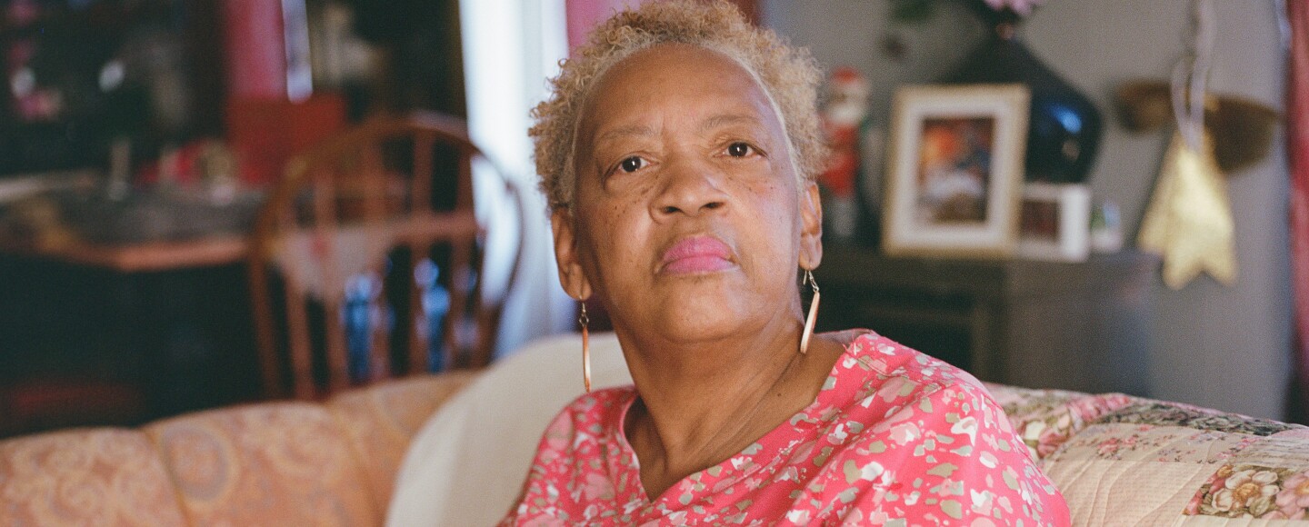 Older Black woman with a pink blouse sitting on a couch and looking straight into the distance