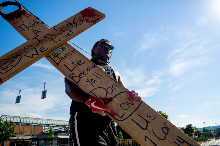 A man carrying a wooden cross also wears a mask, gloves and glasses during a march near Music Hall as people continue to rally and protest the murder of George Floyd as the coronavirus pandemic continues, Wednesday, June 3, 2020, in Cincinnati.