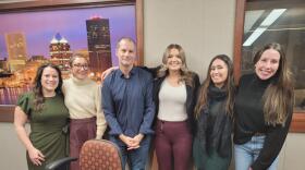 Six people stand with their arms around each other in front of a wall in a radio talk studio: a woman at far left has long brown hair and is wearing an olive green dress; a woman at near left has very short brown hair and is wearing glasses, a beige turtleneck sweater and pink pants; a man to her left has very short dark hair and is wearing a navy button-down shirt and jeans; a woman to his left has long blonde hair and is wearing a black blazer, white shirt and maroon pants; a woman at near right has long brown hair and is wearing a black scarf, a green dress and knee-high black boots; a woman at far right has long brown hair and is wearing a black turtleneck sweater and jeans.