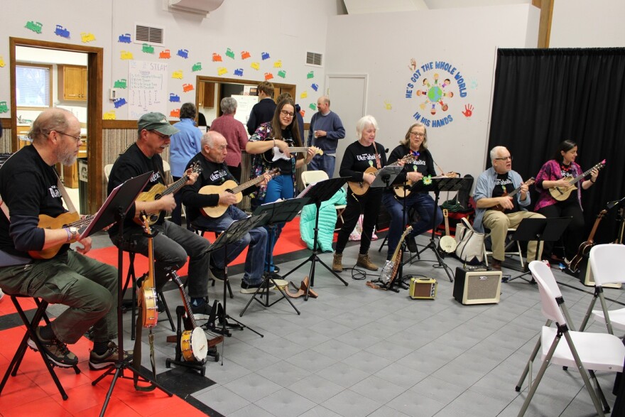 Line of eight people in a semicircle playing ukeleles with a row of people standing behind them with a sign painted on the wall that reads 'he's got the whole world in his hands'