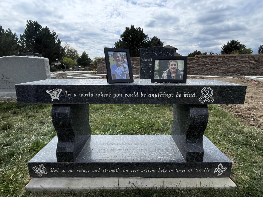 Framed photographs of Alyssa Peterson sit on a memorial bench at her gravesite in Wheat Ridge on Sept. 29, 2026. The bench’s inscription reads, “In a world where you could be anything, be kind.”