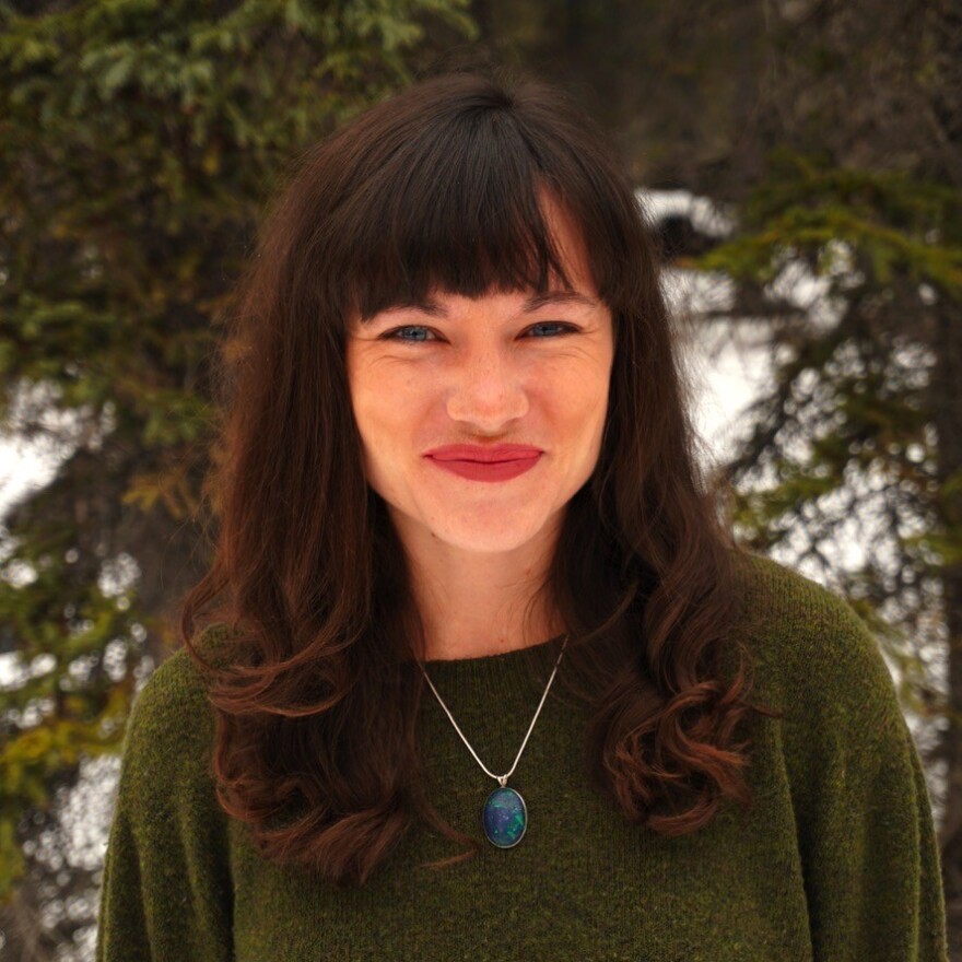 Erin McKinstry is outdoors and smiles at the camera. McKinstry has dark hair and bangs and wears a green sweater and a pendant necklace. The backdrop contains snow and evergreen foliage.