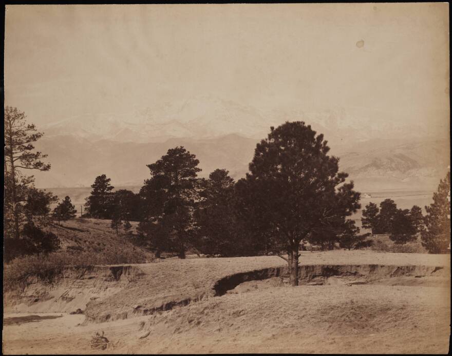 View of foothills and snow on Pikes Peak Austin Bluffs in El Paso County, Colorado. Trees and an eroded wash are on a hillside. A house is in a meadow beside possibly a windmill.