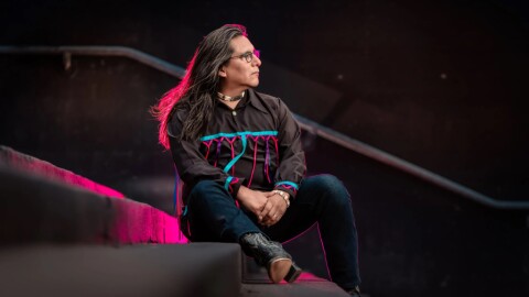 Portrait of Timothy Long wearing a black shirt with colored ribbons, jeans, and hiking boots, sitting on theatre steps