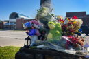 Outdoor photo shows a pile of flowers and stuff animals collecting on top of a concrete pillar. Behind, across the street, is a school building.