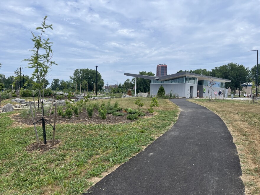 A paved path extends from the foreground to a gray modern building in the back. There are trees and a garden to the left of the path. The M&T tower looms in the background. 