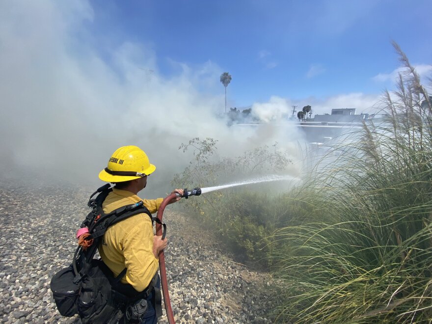 A firefighter holds a hose and sprays water on a smoldering fire in a grassy area.