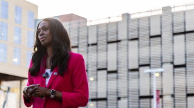 A woman in a pink blazer and a small microphone clipped to her lapel stares ahead.