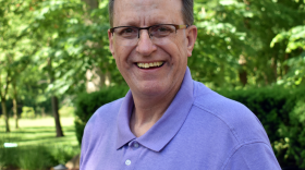  A man wearing glasses smiles at the camera while standing outdoors in front of trees.