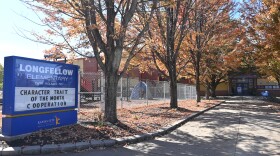 A blue and white school sign sits in the foreground with several trees with orange and brown leaves in front of a brick school building. The sign reads "Longfellow Elementary, Character Trait of the Month: Cooperation."

