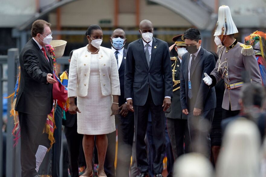 Haiti's president and first lady, both wearing face masks, stand flanked by five men, one of them saluting, outside of an airport.