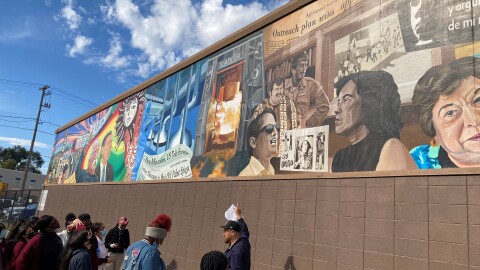 A group of students and a historian gather outside to look at a mural. The mural features many historical scenes in figures in both vivid colors and in sepia tones.