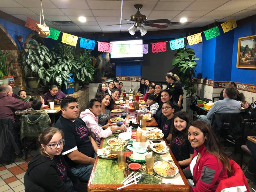 Members of CALY sit along a wooden table at a restaurant.  In the top of the photo a  colorful punched-paper banner hangs.