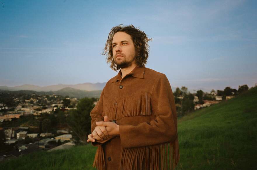 Kevin Morby is dressed in a brown fringed jacket with his hands clasped together staring off in space.