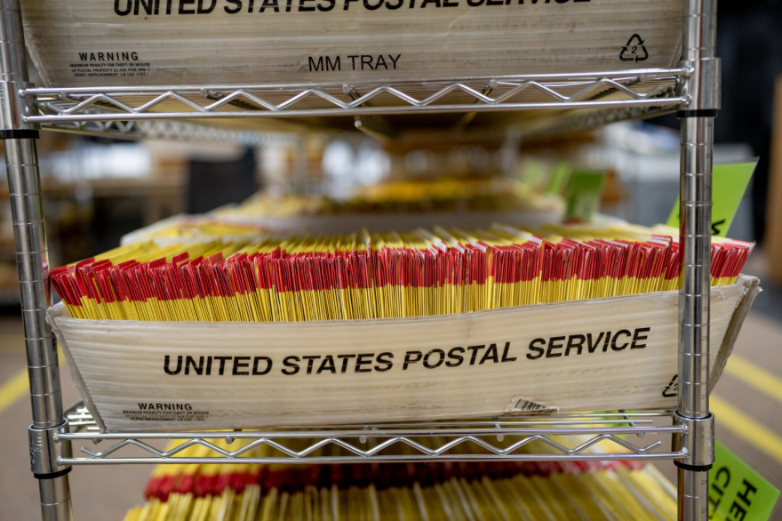 A United States Postal Service-branded tray on a metal shelf stuffed with ballots. Many more trays are partially visible on the same shelf as well as higher and lower shelves.