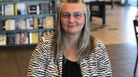 A woman with long graying hair wearing eyeglases sits in what looks like a bookstore with a coffee in front in front of her on the table softly smiling at the camera.