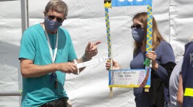 a man in a pale shirt holds a paper needle syringe while his wife looks through a photo frame that says I got vaccinated. 