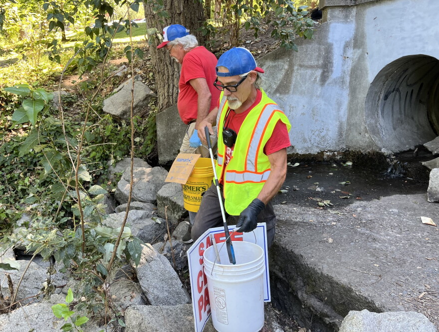 Two middle-aged men in red shirts use trash grabbers to pick up trash in a concrete culvert. 