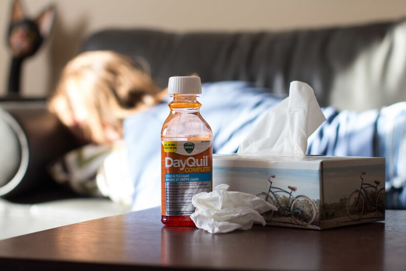 In the fuzzy background a person lays on a couch facing away. In the foreground a bottle of DayQuil and a box of kleenex sit on a brown tabletop.