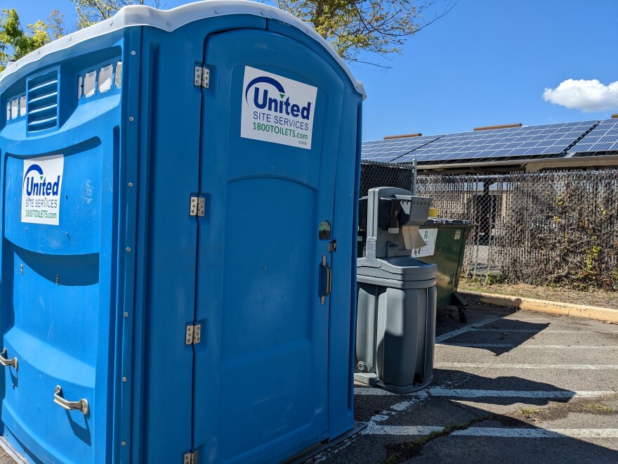 On the left, an accessible portable toilet, a portable handwashing station is just  to the right. And a small metal dumpster is in the background behind the handwashing station.