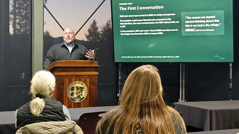 A speaker gestures behind a wooden podium at Oregon Tech beside a presentation slide about reconnecting Lower Klamath Lake and the Klamath River. Audience members sit in the foreground.