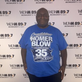 Homer Blow, a long-time radio host in Milwaukee, stands in front of the WUWM 89.7 Milwaukee's NPR photo backdrop wearing a royal blue shirt with the words: "Congratulations Homer Blow Celebrating 35 Years in Radio!"