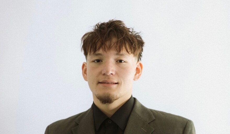 A man in a green button blazer and a black undershirt stands in front of all white background 