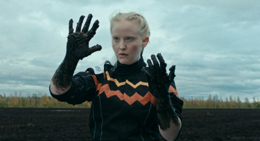 A blond girl with mud on her hands stands in front of a blue sky and muddy field.