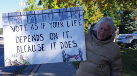 A woman in a tan puffer jacket on Drake Road stands in the shade of a tree holding a white sign that reads in black letters "Vote as if your life depends upon it because it does." 