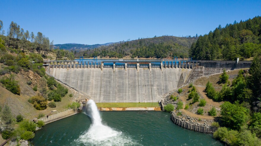 An aerial daytime shot of a concrete dam on a river, facing the reservoir above. some water is spraying at the bottom of the dam into the river below.