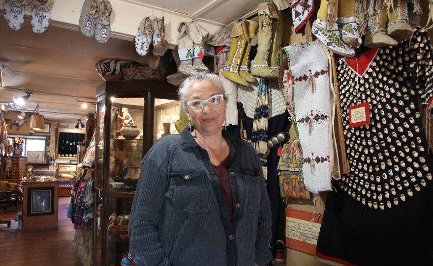 Nico Strange Owl stands and softly smiles at the camera. Behind her is the Indigenous gift shop. There's moccasins hanging from the ceiling, as well as clothing. Directly behind her is a glass case with artifacts.