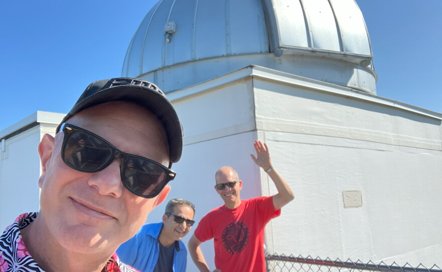  Three men in sunglasses face the camera. The first wears a flowered shirt and a black baseball cap, the second wears a blue shirt and smiles, the third is wearing a red t-shirt and waves. They are all in front of a large rooftop telescope. 