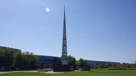 A tall metal spire reaches high into the air, far above the long two story glass building behind it. The spire has metal plates covering it at its base, with a see-through metal mesh covering most of the rest of the structure. The spire's tip is also made with metal plates. A long brown metal wall with WMU's W logo can be seen in front of the structure. 