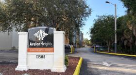 A sign that says "Avalon Heights" in front of the building's driveway. Behind it are the gates and some buildings in the distance.