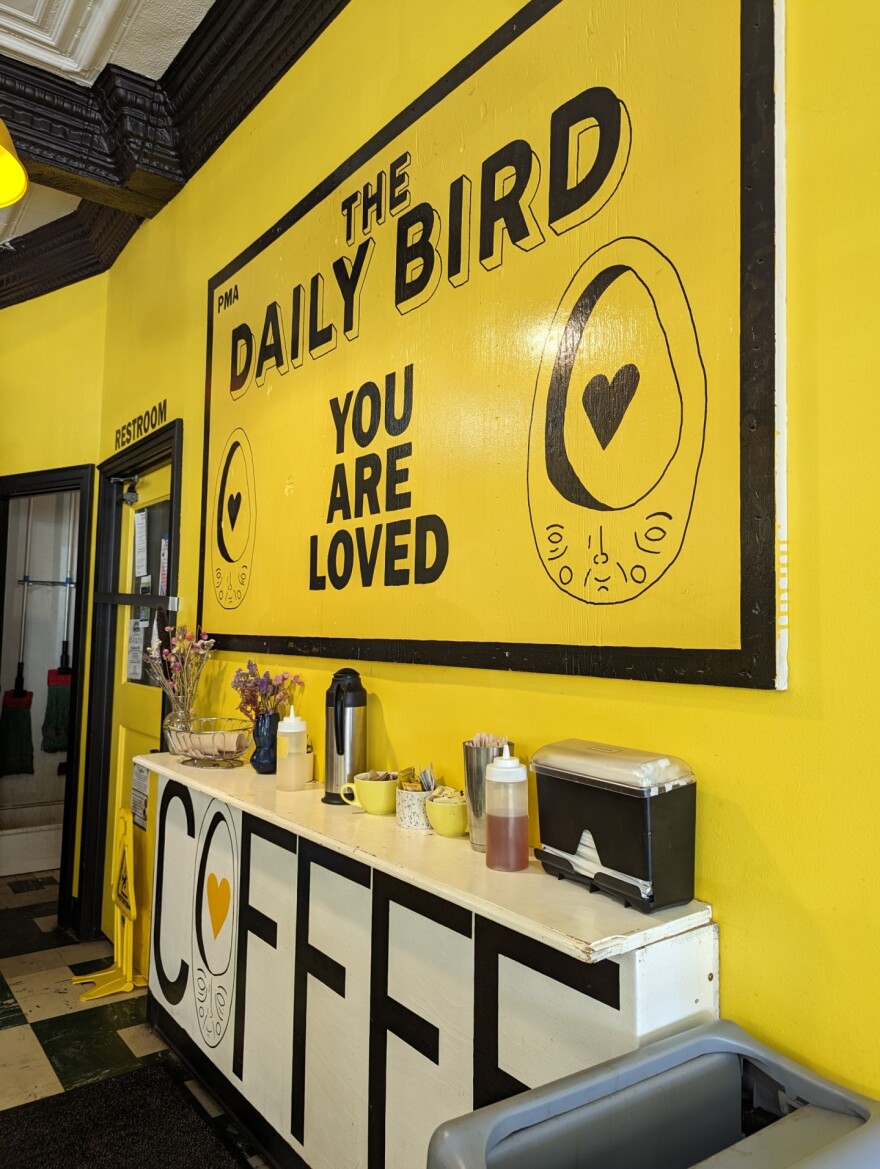 A long table inside a coffee shop with flowers and creamer on top sits against an interior wall painted bright yellow 