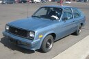 A light Blue three door Chervolet Chevette sits in a parking lot. 