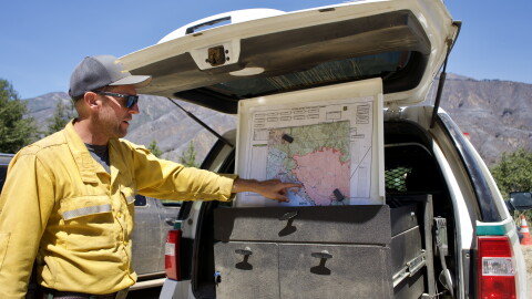 A man in a yellow jacket point  to a map in the back of a truck. 