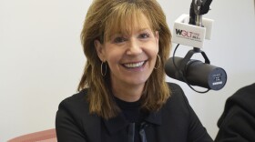 Colleen Kannaday smiling in a radio studio wearing a dark suit and seated next to a microphone with a flag that reads 'WGLT.org 89.1 FM'