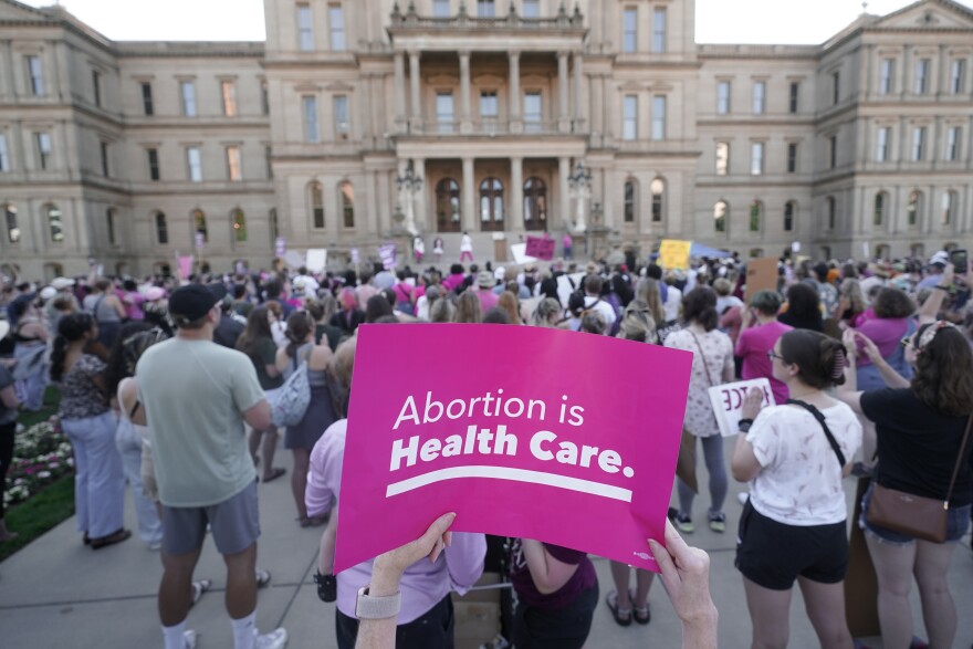A large crowd stands outside the state Capitol. In the foreground is a pink sign that says "Abortion is health care" in white letters.