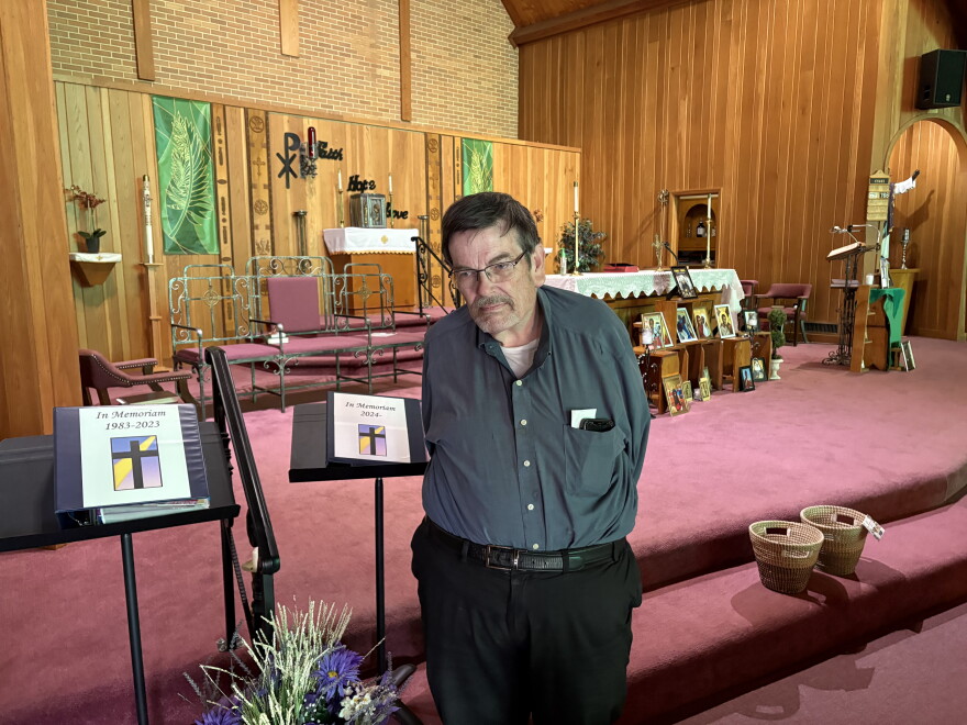 A man stands in front of a church altar. The altar contains a white covering with picture frames in front of it. Music stands are to the left.