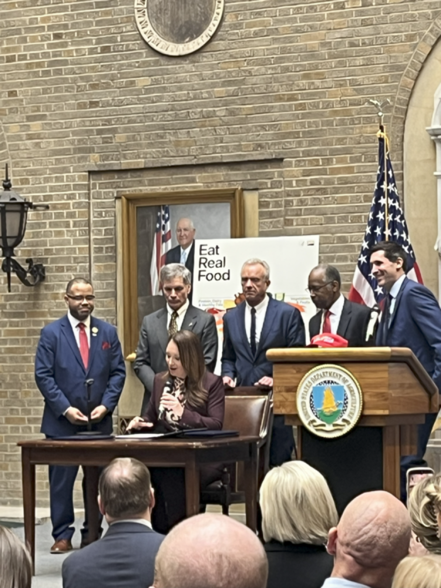 Five men in suits stand behind a woman seated at a desk, who is speaking into a microphone as she looks at a document on the table. Behind them is an American flag and a poster that says, “Eat Real Food” at the top.