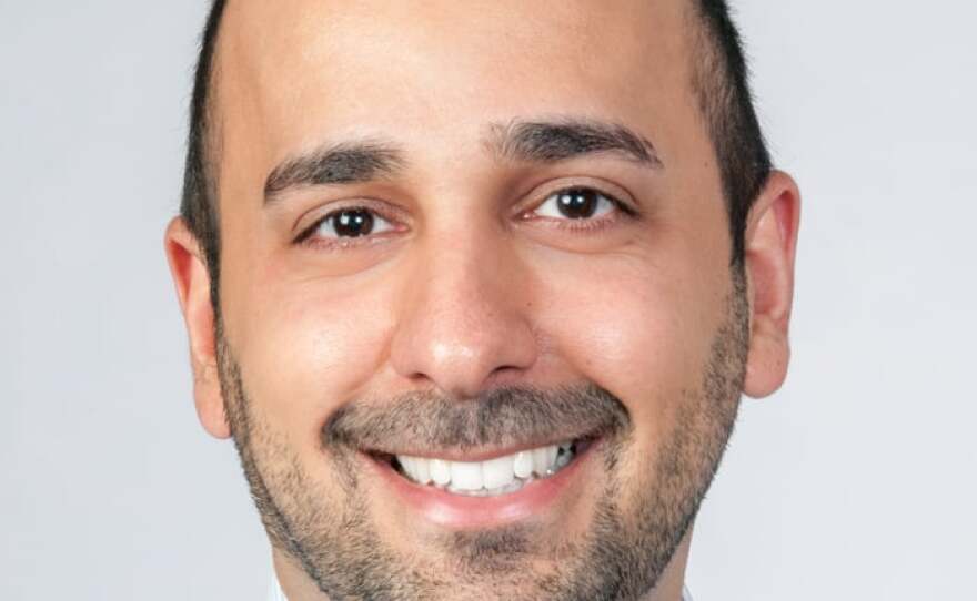 A smiling man with very short dark hair and a dark beard wears a white button-down shirt and blue plaid tie.
