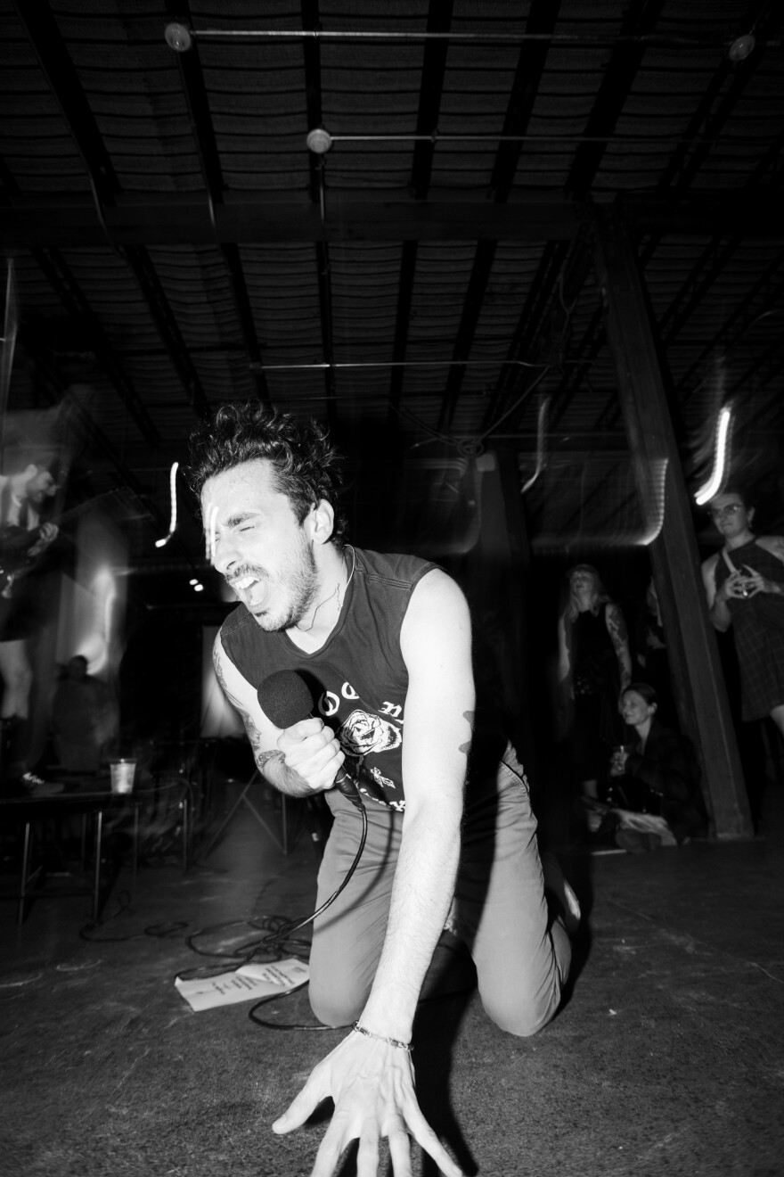 Sal from Gatto Black on his knees, singing passionately into a microphone in a black and white photo.