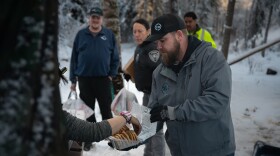 A man in a grey jacket offers a plastic box of cookies to a person out of frame. 